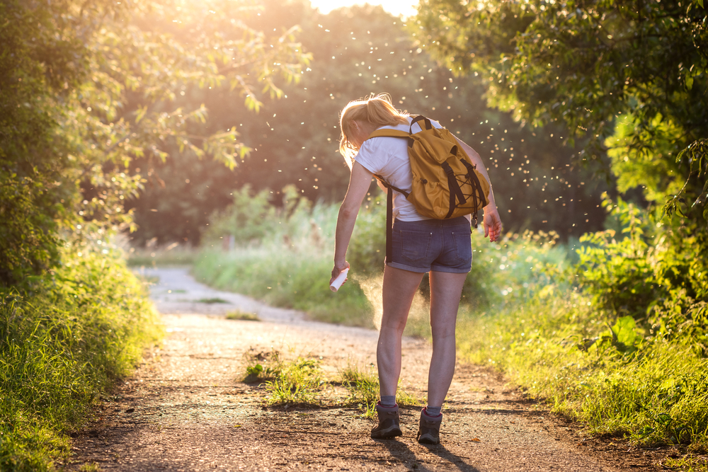 woman standing on path in woods surrounded by insects spraying herself with repellent